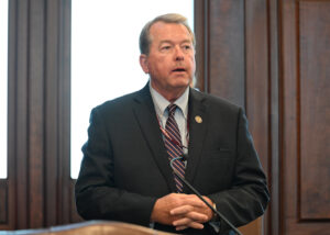 DeWitte at microphone, hands folded